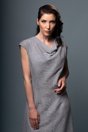 View of the Aurora Aurea draped dress, showcasing its fluid draped neckline, adjustable waist, and soft grey-blue cotton fabric interwoven with gold lurex threads."